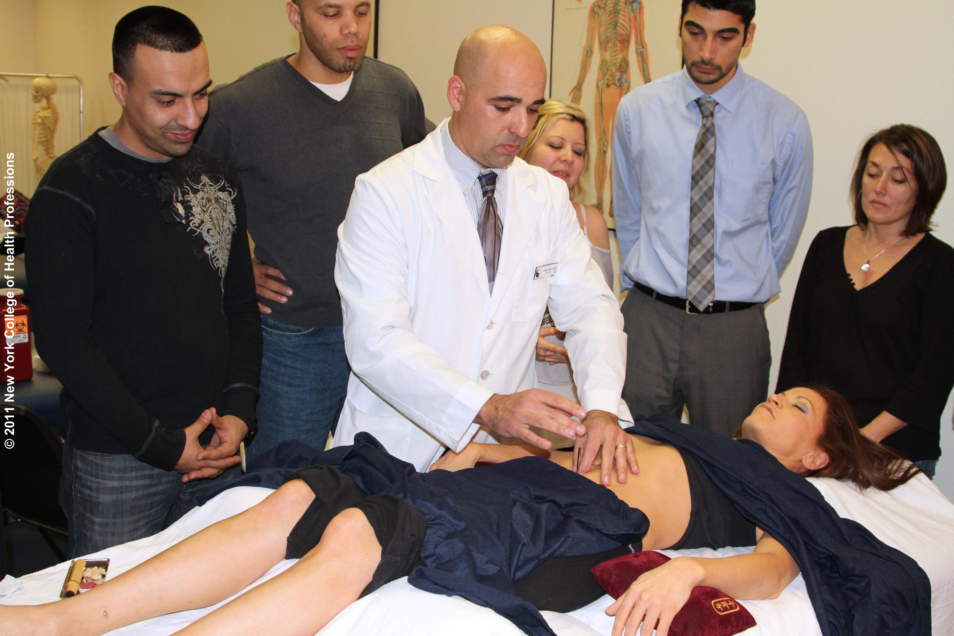A New York College of Health Profession's Japanese Medicine class engaged in the demonstration of an acupuncture procedure. The photo shows an instructor, five students and one person assuming the role of a patient.